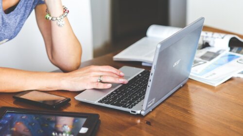Eine Frau arbeitet an ihrem Laptop. Auf dem Tisch liegen auch Magazine, ein Tablet und ein Smartphone